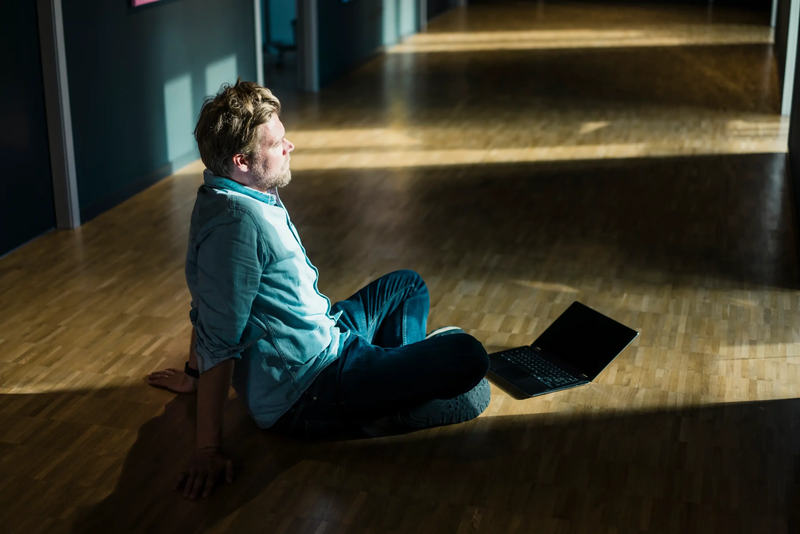 A man sits alone on a wooden floor in a dimly lit hallway, leaning back with a thoughtful expression while looking into a patch of sunlight. A laptop lies open beside him, untouched. The scene conveys deep reflection or regret, visually illustrating the psychological concept of what is hindsight bias—where one reflects on past decisions and feels they should have seen the outcome coming.