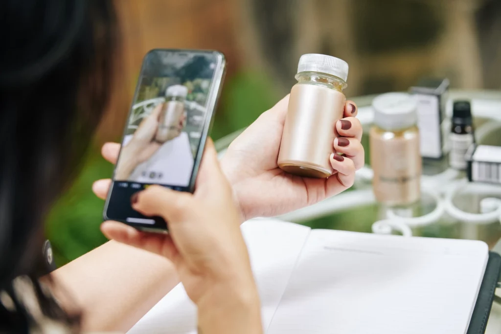 Close-up of a woman using a smartphone to take a photo of a small, metallic skincare or supplement bottle, possibly for product identification or shopping via visual search. The setting includes a blurred background of similar bottles and packaging on a glass table. This moment perfectly illustrates why you should optimize for visual search, as consumers increasingly rely on image-based queries to explore and purchase products instantly.