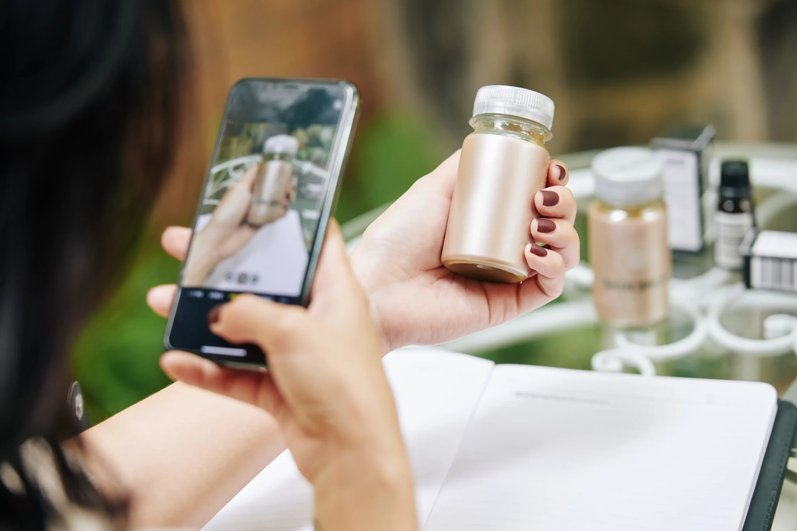 Close-up of a woman using a smartphone to take a photo of a small, metallic skincare or supplement bottle, possibly for product identification or shopping via visual search. The setting includes a blurred background of similar bottles and packaging on a glass table. This moment perfectly illustrates why you should optimize for visual search, as consumers increasingly rely on image-based queries to explore and purchase products instantly.